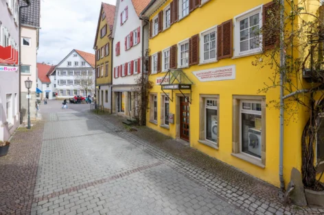 Gasse mit gelbem Gebäude, Schaufenster von Geschäft und einer Buchhandlung auf der linken Seite. Menschen und Autos im Hintergrund.