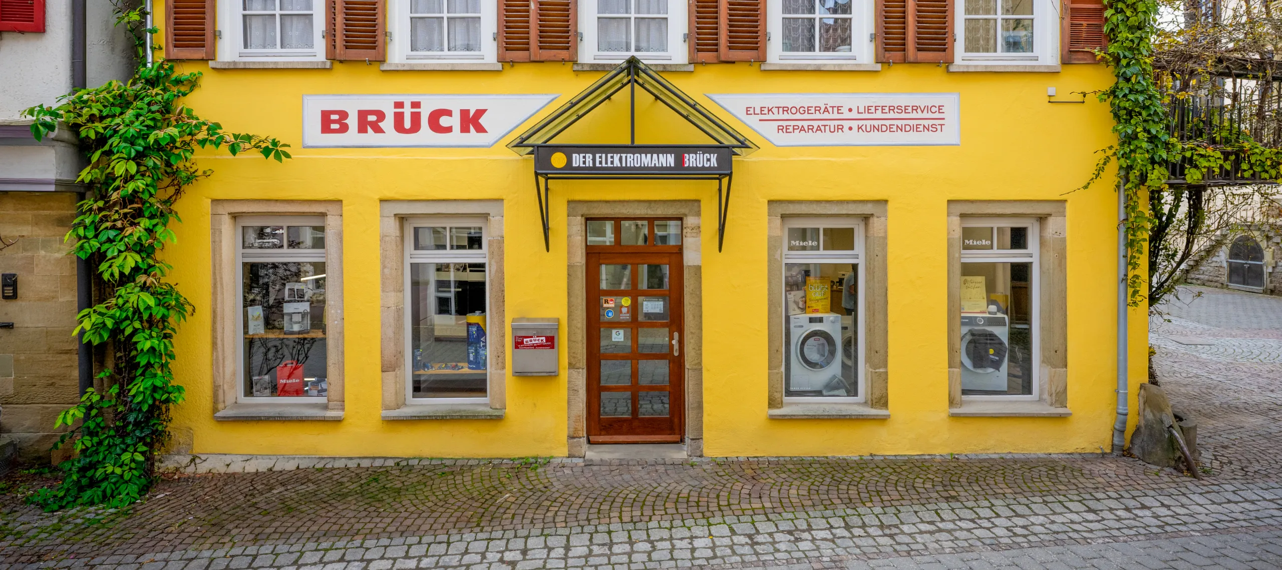 Gelbes Gebäude mit Schaufenstern, Holz-Eingangstür, Schild: BRÜCK, Kletterpflanze, Verkaufsraum mit Haushaltsgeräten sichtbar.