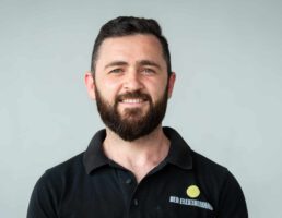 _DSC5419 Mann mit Bart, lächelt, trägt schwarzes Polohemd mit Logo DER ELEKTROMAN. Hintergrund ist grau.