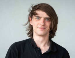 _DSC5516 Junger Mann mit schulterlangem Haar und schwarzen Hemd lächelt in die Kamera, Hintergrund ist schlicht grau.