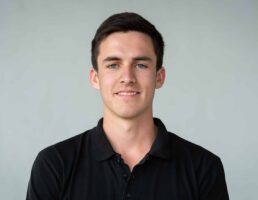 _DSC5585 Junger Mann mit kurzem dunklem Haar und schwarzem Polohemd lächelt leicht vor neutralem Hintergrund.