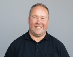 DSC_0268 Mann mit lächelndem Gesicht, grauer Hintergrund, schwarzes Polohemd.