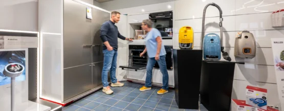 Zwei Personen schauen einen offenen Ofen in einem modernen Küchengeräteladen an; Staubsauger in der Nähe ausgestellt.