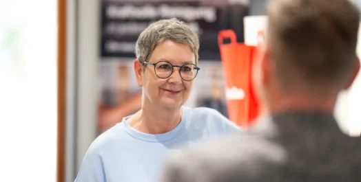 Eine Person mit Kurzhaarschnitt und Brille lächelt, während sie mit einer anderen Person spricht. Hintergrund unscharf.
