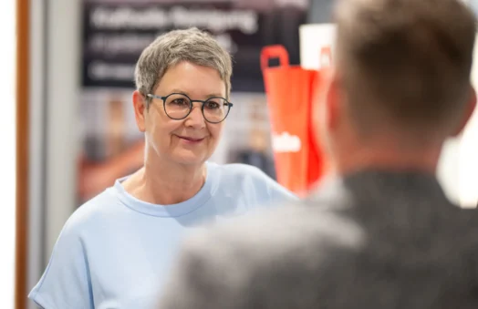 Eine Person mit Kurzhaarschnitt und Brille lächelt, während sie mit einer anderen Person spricht. Hintergrund unscharf.