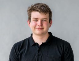 Junger Mann mit kurzem, braunem Haar, trägt ein schwarzes Polo-Shirt, lächelt vor einem grauen Hintergrund.