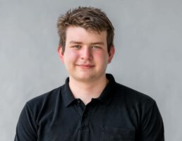 DSC_4988 Junger Mann mit kurzem, braunem Haar, trägt ein schwarzes Polo-Shirt, lächelt vor einem grauen Hintergrund.