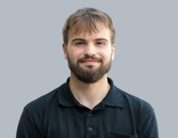 DSC_6981-Bearbeitet Mann mit Bart und kurzem Haar lächelt leicht, trägt ein schwarzes Polohemd vor grauem Hintergrund.