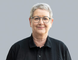 Person mit kurzem grauen Haar, runder Brille, schwarzem Hemd, lächelt freundlich vor grauem Hintergrund.