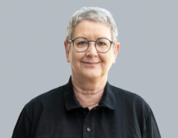 DSC_7130-Bearbeitet Person mit kurzem grauen Haar, runder Brille, schwarzem Hemd, lächelt freundlich vor grauem Hintergrund.