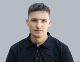 DSC_7169-Bearbeitet Ein junger Mann mit kurzem Haar, schwarz gekleidet, schaut ernst in die Kamera. Grauer Hintergrund.