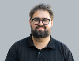 DSC_7219-Bearbeitet Mann mit Brille und Bart in schwarzem Hemd vor grauem Hintergrund.