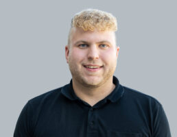 DSC_7301-Bearbeitet Mann mit lockigem, blondem Haar und Bart, trägt schwarzes Poloshirt, lächelt vor grauem Hintergrund.