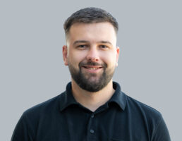 DSC_7374-Bearbeitet Lächelnder Mann mit Kurzhaarfrisur und Bart, trägt ein schwarzes Poloshirt, grauer Hintergrund.