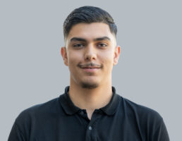 DSC_7434-Bearbeitet Mann mit kurzem, dunklem Haar und Bart trägt ein schwarzes Polohemd vor grauem Hintergrund, lächelt leicht.