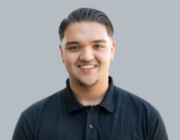 DSC_7539-Bearbeitet Junger Mann mit Kurzhaarschnitt und schwarzem Poloshirt, lächelt vor grauem Hintergrund.