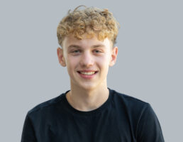 DSC_7601-Bearbeitet Lächelnder junger Mann mit lockigem Haar trägt ein schwarzes T-Shirt vor grauem Hintergrund.