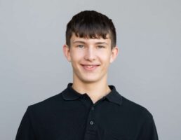 Junger Mann mit kurzem, dunklem Haar und freundlichem Lächeln, trägt ein schwarzes Poloshirt, vor grauem Hintergrund.