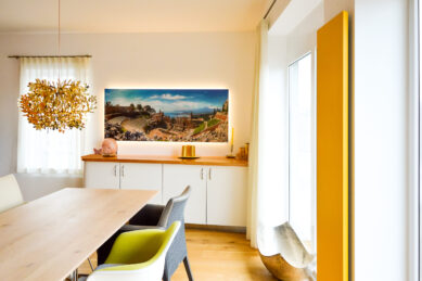 Esszimmer mit Tisch, bunten Stühlen, dekorativem Kronleuchter, großem Landschaftsbild und Fenster mit gelbem Rahmen.