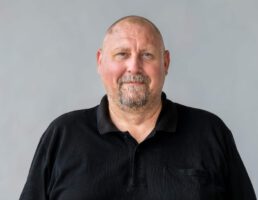 Ein bärtiger Mann mit Glatze trägt ein schwarzes Polohemd. Er steht vor einem hellgrauen Hintergrund und lächelt leicht.