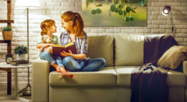 Frau und Kind auf Sofa, lesen Buch, Lampenlicht, helles Wohnzimmer, Kuscheltier, Pflanzen, Bild von Oliven an der Wand.
