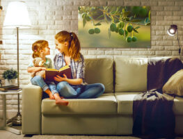 Frau und Kind auf Sofa, lesen Buch, Lampenlicht, helles Wohnzimmer, Kuscheltier, Pflanzen, Bild von Oliven an der Wand.